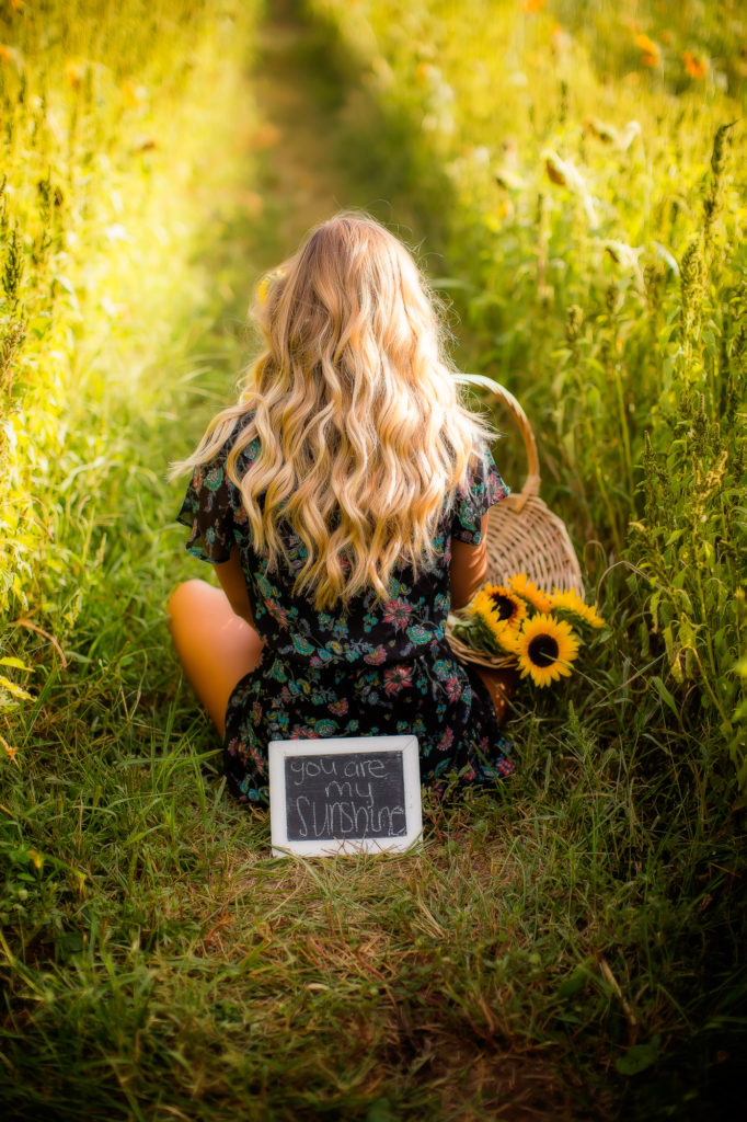 Girl sitting chris-cross-applesauce holding a basket of sunflowers with a sign placed behind her that says "you are my sunshine"