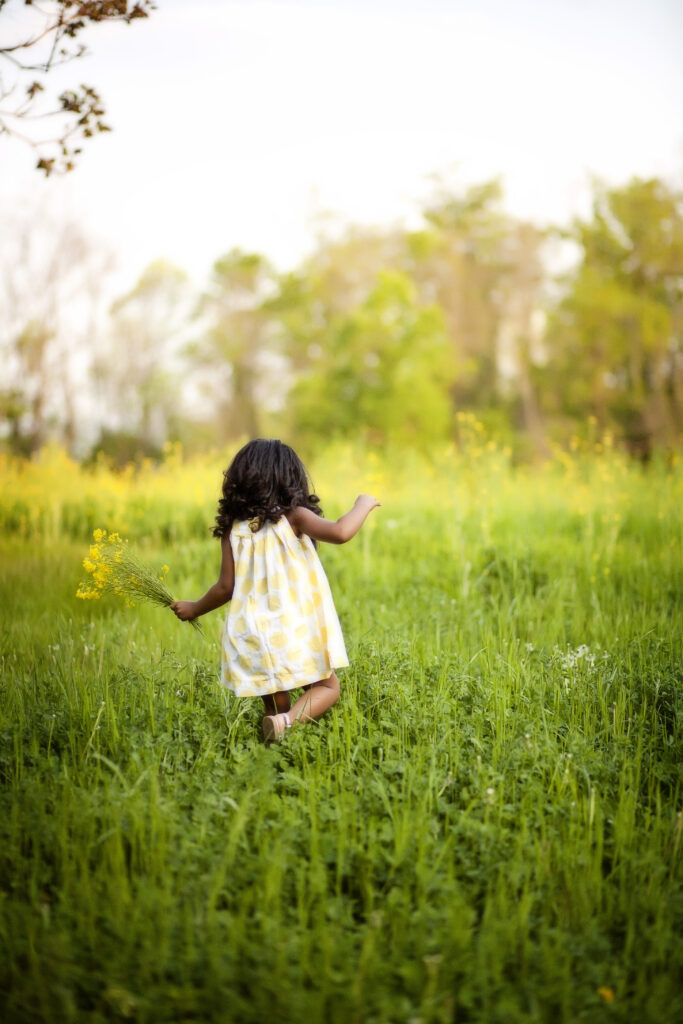 Three year old little girl holding wild flowers in a yellow sun dress walking away from the camera in a field.