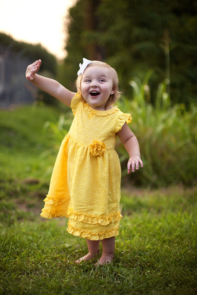 Four year old girl in a yellow dress with her right hand up smiling. 