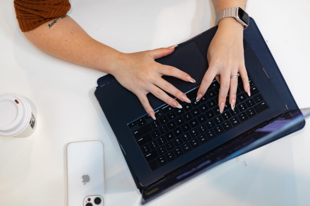 Female hands with painted nails typing on a laptop with her iphone sitting next to her and a cup of coffee.