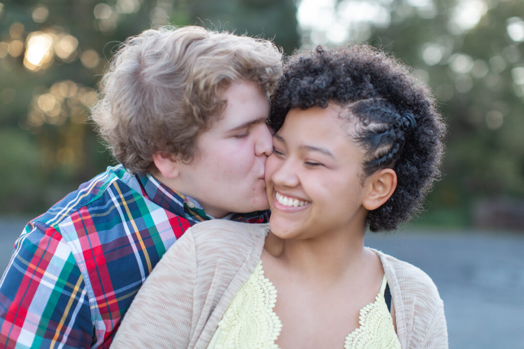 Teenage couple with the male hugging the female from behind and kissing her on her smiling cheek.