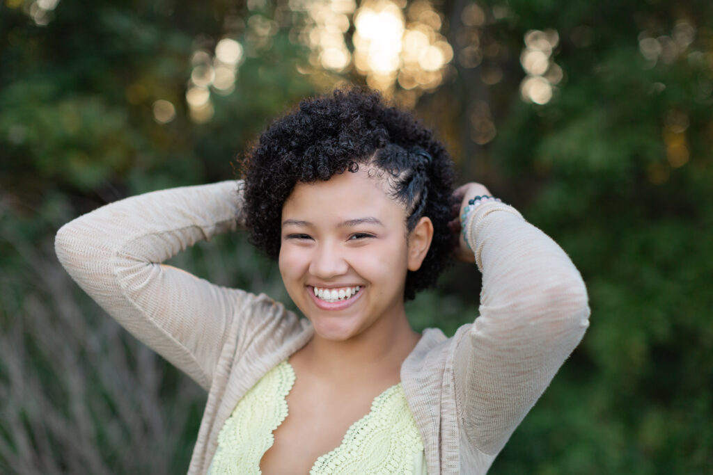 Female with her hands behind her head with a yellow shirt and tan sweater on smiling.  Senior portrait session planning